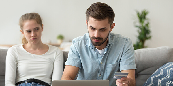 Husband paying bill online with wife in background with displeased face
