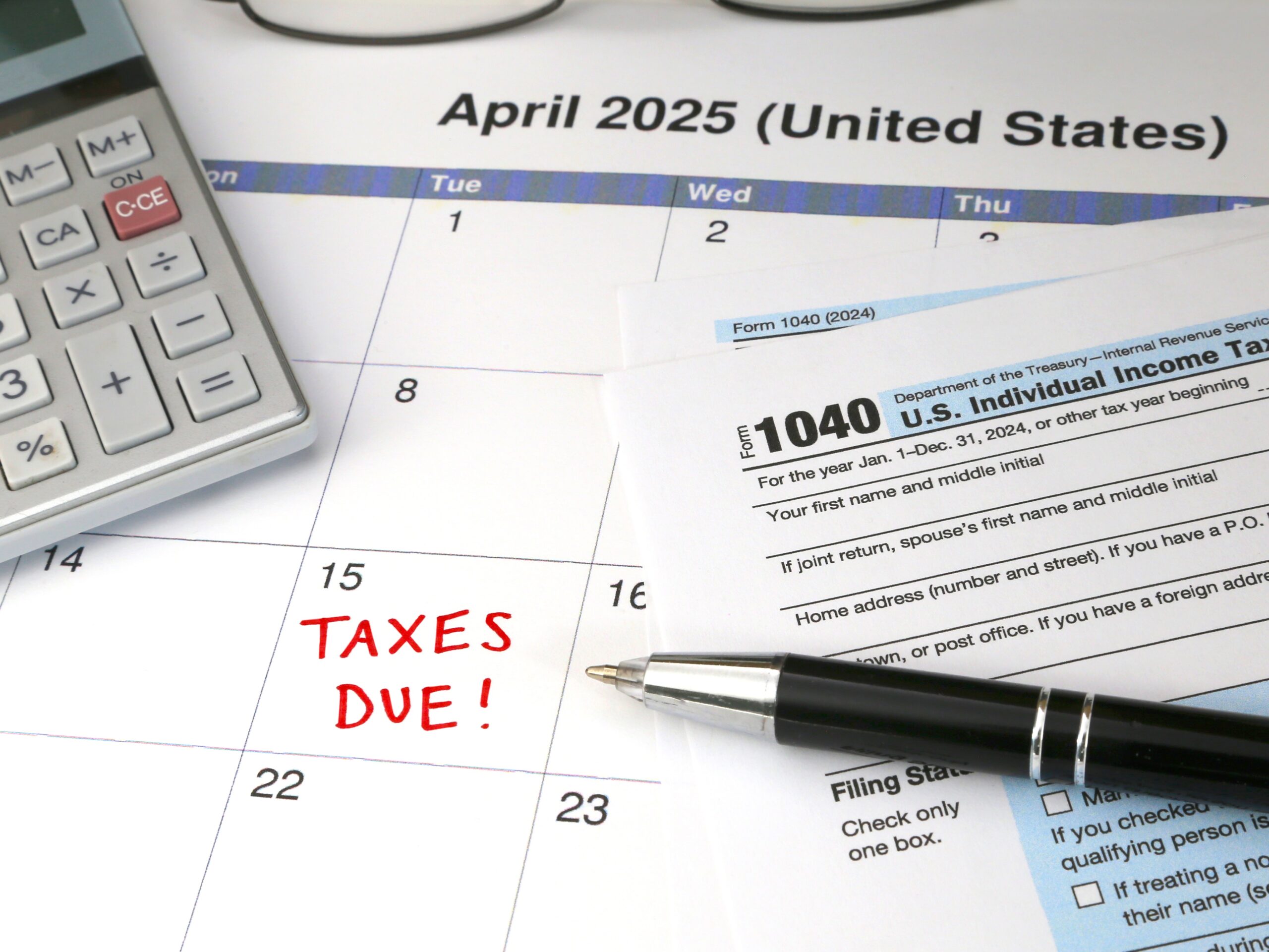 A 2025 calendar noting the April 15 USA Internal Revenue Service IRS income filing deadline for year 2024 taxes is shown up close, with a calculator, ink pen, and glasses in the frame.