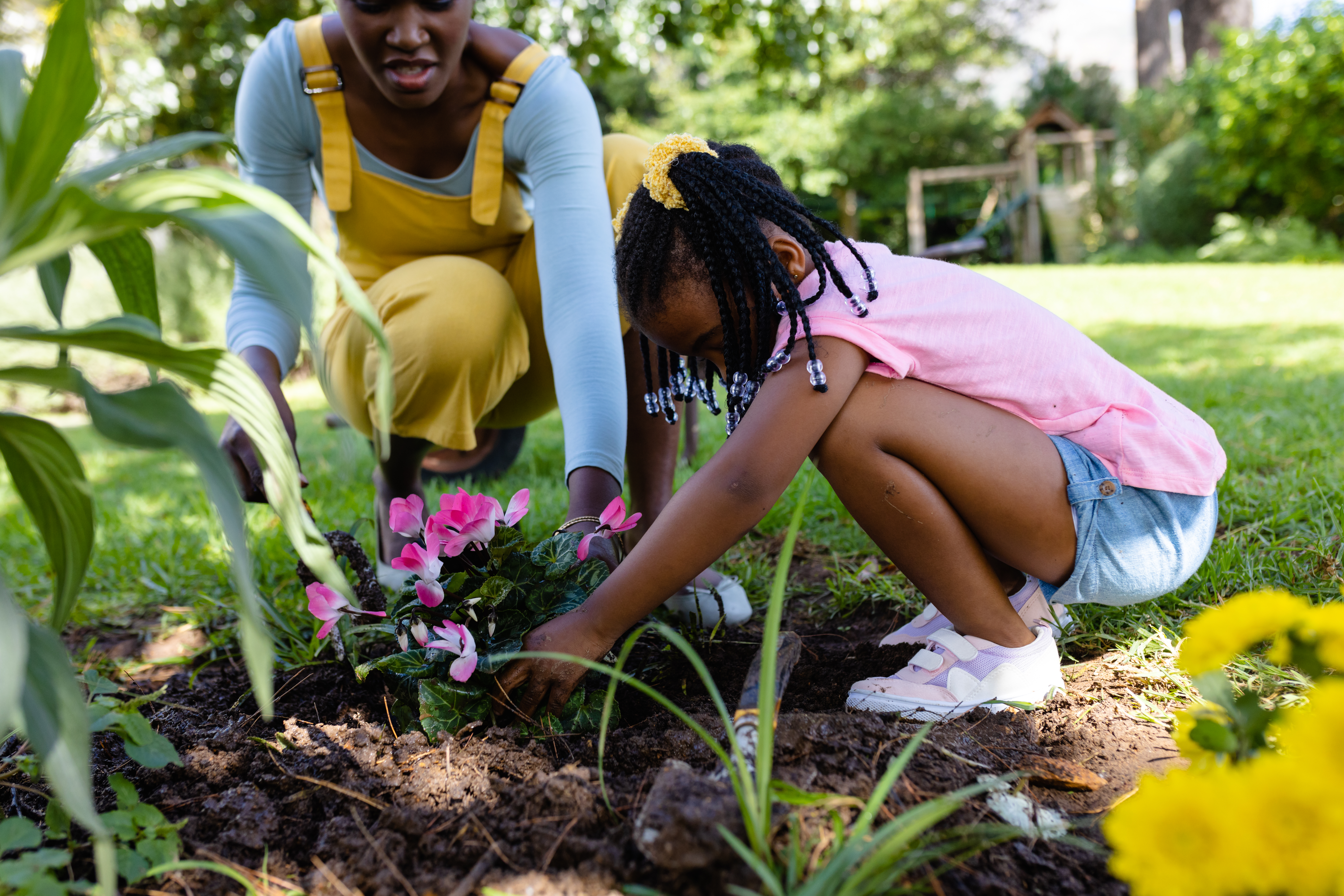 A woman and a child plant pink flowers in a garden. The child, wearing a pink shirt, focuses intently. It's a bright, sunny day.